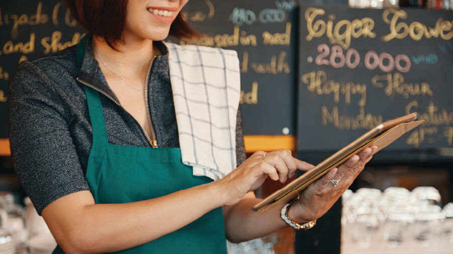 A close-up of a cafe staff member wearing a green apron, using a finger to operate an iPad tablet in front of a chalkboard menu, illustrating how to choose stable bluetooth hardware and accessories for an eRunlite iPad POS system.