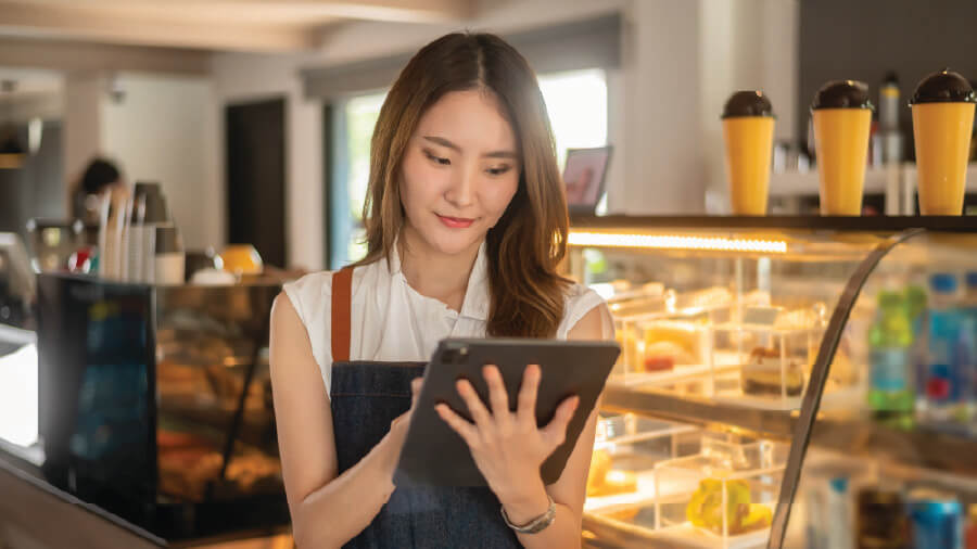 A smiling young female bakery owner holding a tablet to manage order billing next to a pastry display case, representing the easy-to-use and unified interface of eRunlite tablet POS system recommended for modern cafes and retail shops.