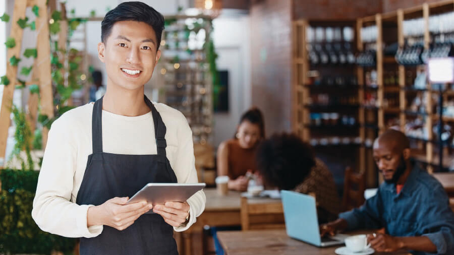 A smiling cafe server wearing an apron and holding a tablet in a busy coffee shop background, representing shop owners upgrading from a traditional cash register to the eRunlite cloud POS system.